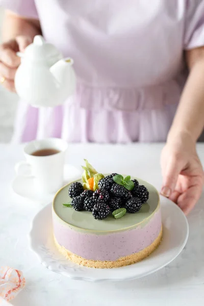 軽めのテーブルの上には、抹茶とホワイトチョコレートとベリーのココナッツチーズケーキ。女の子の手がやかんを握っている. ロイヤリティフリーのストック写真