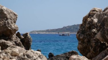 Türk balıkçı teknesi denizin açık sularında yelken açıyor. Parlak güneşli bir gün. Sahildeki taşlar ön planda. Suda yaz yürüyüşü..