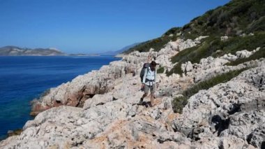 Büyük sırt çantalı bir kız turist güneşli bir günde, denizin yakınındaki dağlık bir bölgede, keskin kayalar boyunca ihtiyatlı bir şekilde yürür..