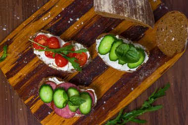 Three kinds of sandwiches with cottage cheese and vegetables: cucumbers, tomatoes, basil and arugula on dark bread dusted with flour on a wooden kitchen dividing board. For websites, menus, advertising banners on the subject of cooking, kitchen, food
