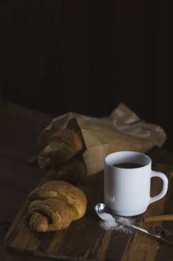 Baked fresh croissants with chocolate in the kitchen interior on a wooden dividing board on a dark background. Nearby is a white cup of coffee with sugar and cinnamon. Photos of food for cafes, restaurants, home kitchens and other culinary topics.