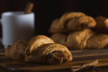 Baked fresh croissants with chocolate in the kitchen interior on a wooden dividing board on a dark background. Nearby is a white cup of coffee with sugar and cinnamon. Photos of food for cafes, restaurants, home kitchens and other culinary topics.