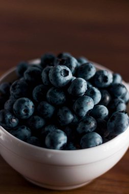 A handful of fresh blueberries in a white deep plate with handles on a wooden kitchen table in a dark modern interior. Topics of food, products, vitamins, health, cooking