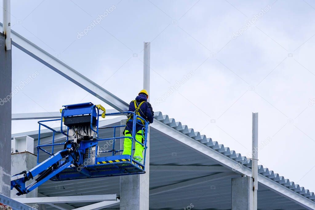 a worker in uniform and safety protective equipment using a self propelled elevator assembles the metal structures of a building