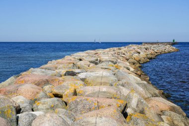A breakwater of large stones stretches into the Baltic Sea, clear blue sky to the horizon