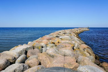 A breakwater of large stones stretches into the Baltic Sea, clear blue sky to the horizon