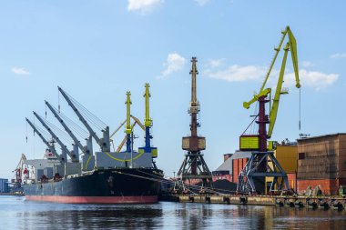 Liman rıhtımında açık ambarı olan büyük bir gemi, kıyı vinçleri ve kömür yığınları, mavi gökyüzü arka planı