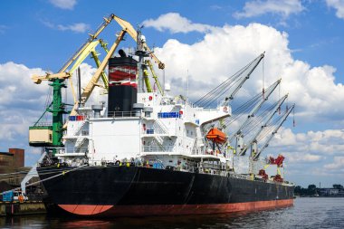 Liman kargo terminalinde açık ambarı olan büyük bir gemi, kıyı vincleri, mavi gökyüzü arka planı