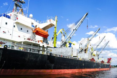 Liman kargo terminalinde açık ambarı olan büyük bir gemi, kıyı vincleri, mavi gökyüzü arka planı