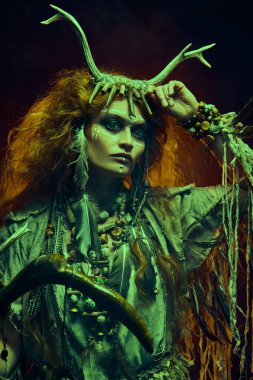 Şaman kadın portresi, ayin kıyafetleri ve geyik boynuzu başlığıyla arkasında alevlerle yanan karanlık arka planda poz veriyor. Etnik gelenekler. Paganizm. Cadılar Bayramı.