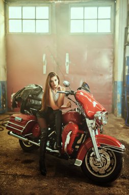 Diz kapağında siyah deri, deri şort ve üstü eski bir garajda motosikletinin üzerinde oturan kendine güvenen güzel bir motorcu kız. Tam boy portre.