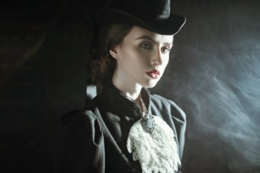 19. yüzyıldan kalma takım elbiseli zarif genç bayan dumanla dolu karanlık bir odada poz veriyor. Tarihsel saç stili ve makyaj.