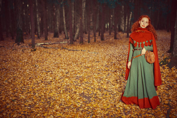 Full length portrait of a young red-haired girl of the Middle Ages walking through a dense autumn forest. Ancient times. Celts. Fantasy world. Copy space.
