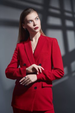 Zarif bir iş tarzı. Zarif makyajlı güzel bir genç kadının portresi. Koyu gri arka planda gölgeli, zarif kırmızı bir takım elbiseyle poz veriyor. Kadınlık, feminizm ve moda.
