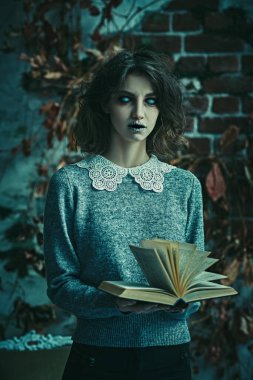Spooky portrait of a poltergeist girl with colorless eyes standing in a gloomy room by a brick wall with a mysterious book in her hands. Halloween. Horror scene.  clipart