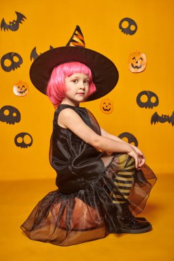 Cadı kostümü giymiş komik küçük kız balkabağının üzerinde oturuyor. Cadılar Bayramı süslemeleriyle dolu sarı bir arka planda tam boy portre. Cadılar Bayramı. 