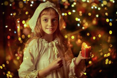 Gecelikli, şapkalı, neşeli küçük bir kız Noel gecesi elinde mumla neşeyle gülümsüyor. Sihir zamanı. Eski günlerdeki Noel. 