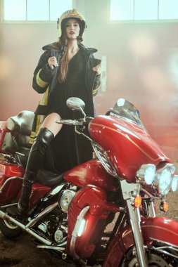 Garajda kırmızı motosikletle poz veren itfaiyeci gibi giyinmiş çekici bir kız..