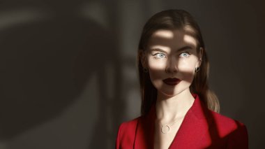 Zarif bir iş tarzı. Zarif makyajlı güzel bir genç kadının portresi. Yüzünde gölgeler olan koyu gri bir arka planda zarif kırmızı bir takım elbiseyle poz veriyor. Kadınlık, feminizm ve moda.