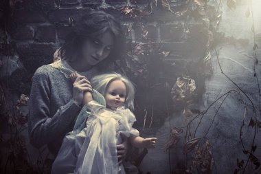 Ürkütücü, solgun gözlü bir hayalet kadın yıkılmış bir duvarın yanında solmuş yaprakların arasında duran bir bebekle gülümsüyor. Korku filmi, gerilim. Cadılar Bayramı.