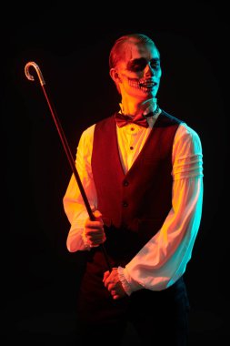 Kafatası makyajlı zarif giyimli genç bir adamın portresi elinde bastonla siyah bir arka planda gülümsüyor. Ürkütücü kırmızı ışık. Dia de los muertos. Ölüler Günü. Cadılar Bayramı.