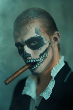 Zarif takım elbiseli, yüzünde kafatası makyajı olan ve ağzında puroyla poz veren şeytani yakışıklı bir adam. Dia de los muertos. Ölüler Günü. Cadılar Bayramı.