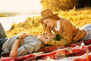 Romantik genç çift, güneşli bir günde, nehrin kenarındaki battaniyenin üzerinde uzanarak harika bir günün tadını çıkarıyor. Aşk konsepti. Sonbahar mevsimi.
