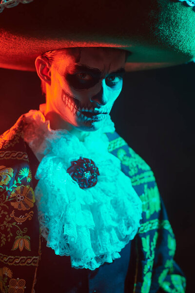 Портрет красивого элегантного мужчины в мексиканском сомбреро и традиционном костюме с жутким макияжем черепа на лице. Черный фон с красным светом. Dia de los muertos. День Мертвых. Хэллоуин.