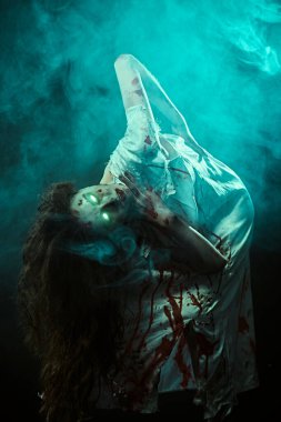 Korkunç şekilsiz, yanan gözlü bir kadın, şeytan tarafından ele geçirilmiş, kanlı bir gecelik kıvrılmış, karanlıkta duruyor, dumanla çevrili. Ürkütücü zombi kadın. Korku, gerilim. Cadılar Bayramı.