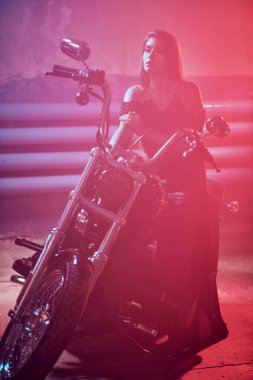 Siyah bir gece elbisesi içindeki çarpıcı genç kadın garajda parlak kırmızı ışıkla aydınlatılmış bir motosikletin yanında poz veriyor. Moda fotoğrafı..