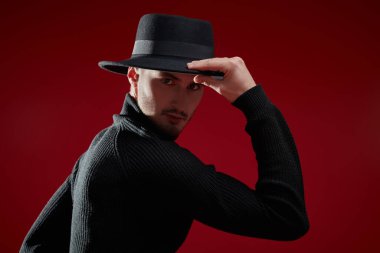 Erkek modası ve tarzı. Yakışıklı esmer adam siyah balıkçı yaka ve şapkayla poz veriyor. Kırmızı stüdyo arka planı. 