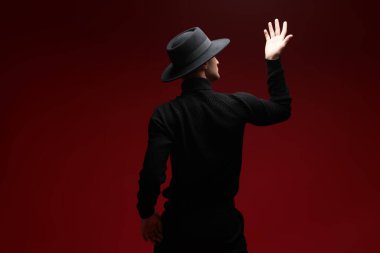 Model bir adamın portresi, şık siyah günlük kıyafetler ve kameraya arkası dönük poz veren bir şapka. Kırmızı stüdyo geçmişi. Erkek stili ve modası. 