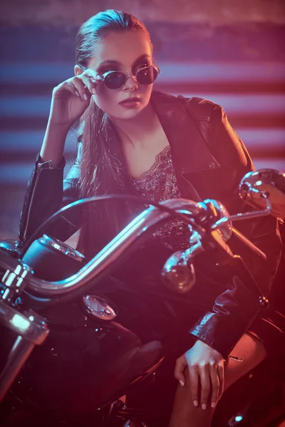 Confident girl biker in black leather jacket and stylish black sunglasses poses sitting on her motorcycle in a garage.