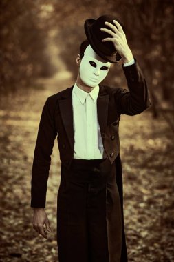 Beyaz karnaval maskeli ve siyah ceketli gizemli bir beyefendi melon şapkasını kaldırarak eski bir sonbahar parkında duruyor. Pandomim, karnaval. Cadılar Bayramı. Klasik stil.