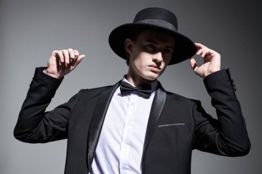 Gri bir stüdyo arka planında poz veren şık siyah takım elbiseli, papyonlu ve şapkalı bir mankenin portresi. Erkek modası ve güzelliği. 