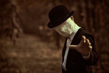 Yüzünde alçı bandajlı, melon şapkalı ve siyah ceketli korkunç bir beyefendi, eski bir sonbahar parkında duruyor ve elini uzatıyor. Cadılar Bayramı. Eski tarz. Korku, gerilim.