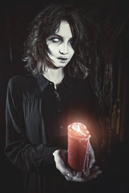 A lonely ghost girl in a black mourning dress stands with a candle in her hands in a dark gloomy room, shrouded in cobwebs. Horror film, thriller. Halloween. clipart