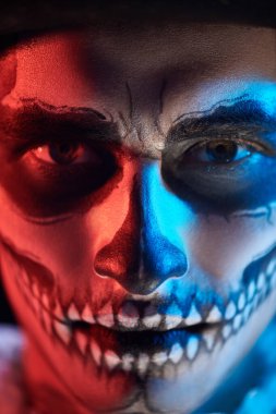 Kafatası makyajlı, koyu kırmızı ve mavi ışıklı şeytani yakışıklı bir adamın yakın plan portresi. Dia de los muertos. Ölüler Günü. Cadılar Bayramı.
