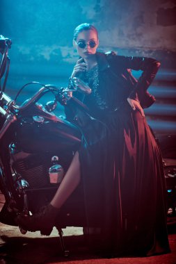 Siyah pullu gece elbisesi ve deri ceketiyle muhteşem bir manken garajda kırmızı ve mavi ışıkla aydınlatılmış bir motosiklet üzerinde poz veriyor. Moda fotoğrafı..