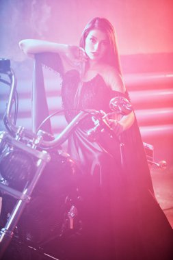 Uzun siyah bir gece elbisesi içindeki çarpıcı genç kadın garajda parlak kırmızı ışıkla aydınlatılmış bir motosikletin yanında poz veriyor. Moda fotoğrafı..