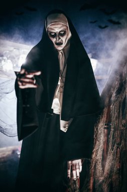 Cadılar Bayramı. Şeytani lanetli rahibe elini ileri uzatıyor, terk edilmiş kilisede duruyor, ürkütücü bir sis ile çevrili. Dehşet.