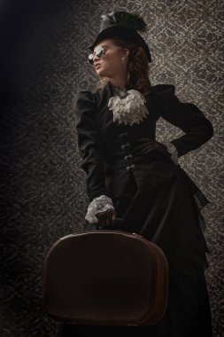 Geleneksel 19. yüzyıl kıyafetleri ve yuvarlak gözlüklü zarif bir kadının portresi. Eski bir arka planda ellerinde seyahat çantasıyla poz veriyor. 19. yüzyılın sonları - 20. yüzyıl başları.