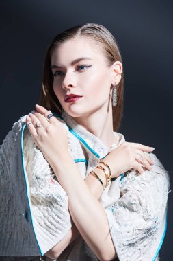 Moda çekimi. Zarif makyajlı güzel bir manken siyah bir arka planda takılar ve geniş kollu abartılı bir bej bluz giyiyor. Stüdyo resmi.