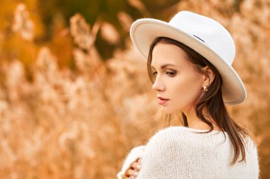 Beyaz yün kazaklı ve beyaz keçeli şapkalı zarif genç kadın sonbahar parkında sazlıkların arkasında poz veriyor. Sonbahar modası ve güzelliği. Boşluğu kopyala.