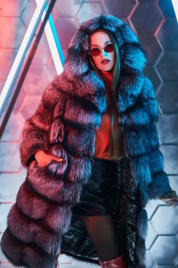 Pahalı gümüş tilki kürklü güzel bir manken kız ve neon lambaların ışığında poz veren şık güneş gözlükleri. Şık kürk manto modası. Tekno biçim.