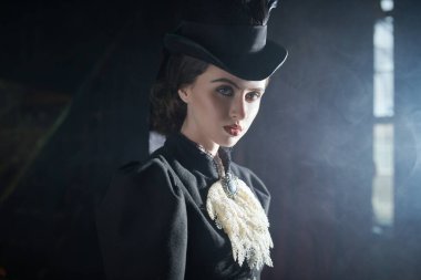 19. yüzyıldan kalma takım elbiseli zarif genç bayan dumanla dolu karanlık bir odada poz veriyor. Tarihsel saç stili ve makyaj.