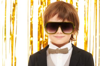 Zarif bir gece elbisesi ve parlak altın folyo perdelerle arka planda poz veren şık güneş gözlüklü şirin bir çocuğun portresi. Çocuk modası ve tatiller. Çocuk partisi..