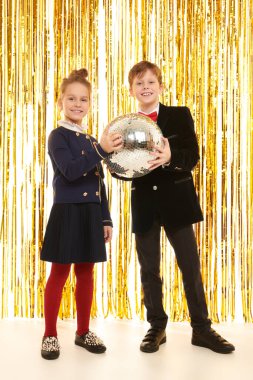 Çocuk partisi. Gece kıyafetleri içinde neşeli küçük bir kız ve oğlan, arka plandaki parlak altın folyo perdelerde bir disko topuyla neşeli bir partide poz veriyorlar. Çocuk modası ve tatiller. 