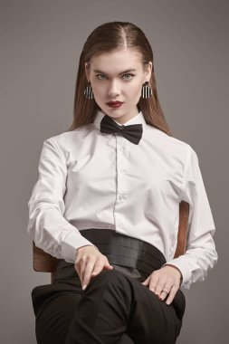 Beyaz gömlekli, papyonlu, siyah pantolonlu, parlak makyajlı genç bir kadın gri arka planda bir sandalyeye oturmuş poz veriyor. İş stili. Başarılı insanlarız. Moda çekimi.. 