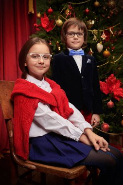 Zarif klasik okul üniforması ve şık gözlüklü iki modern çocuk bir Noel ağacının yanında lüks bir iç mekanda poz veriyorlar. Noel süsü. Çocukların okul modası..